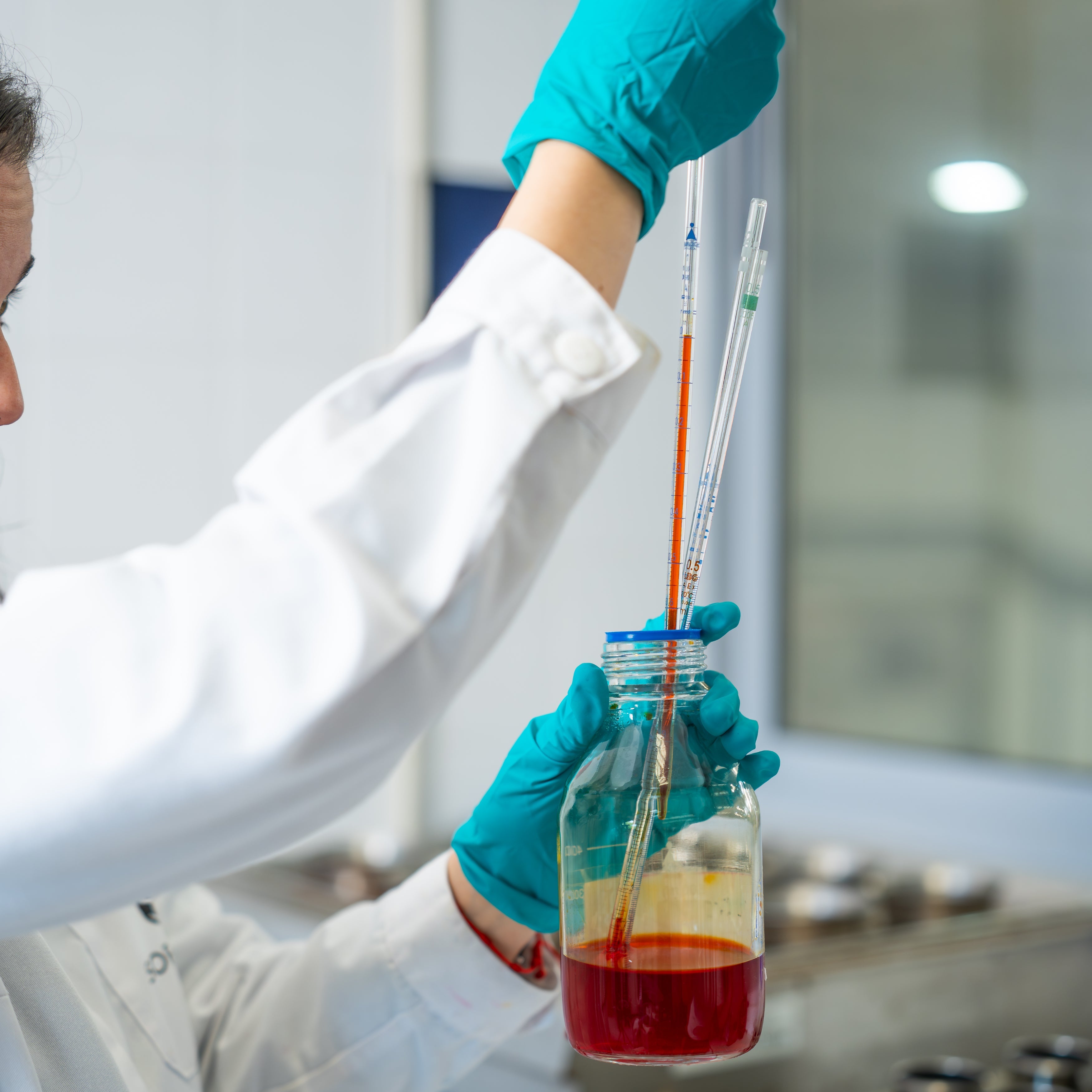 Person in a lab coat and gloves holding a bottle with a red liquid and a pipette.